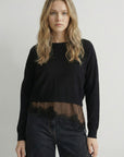 A woman modeling a black long-sleeve knit top with a sheer brown mesh band at the hem, paired with dark jeans, against a plain light gray studio background.