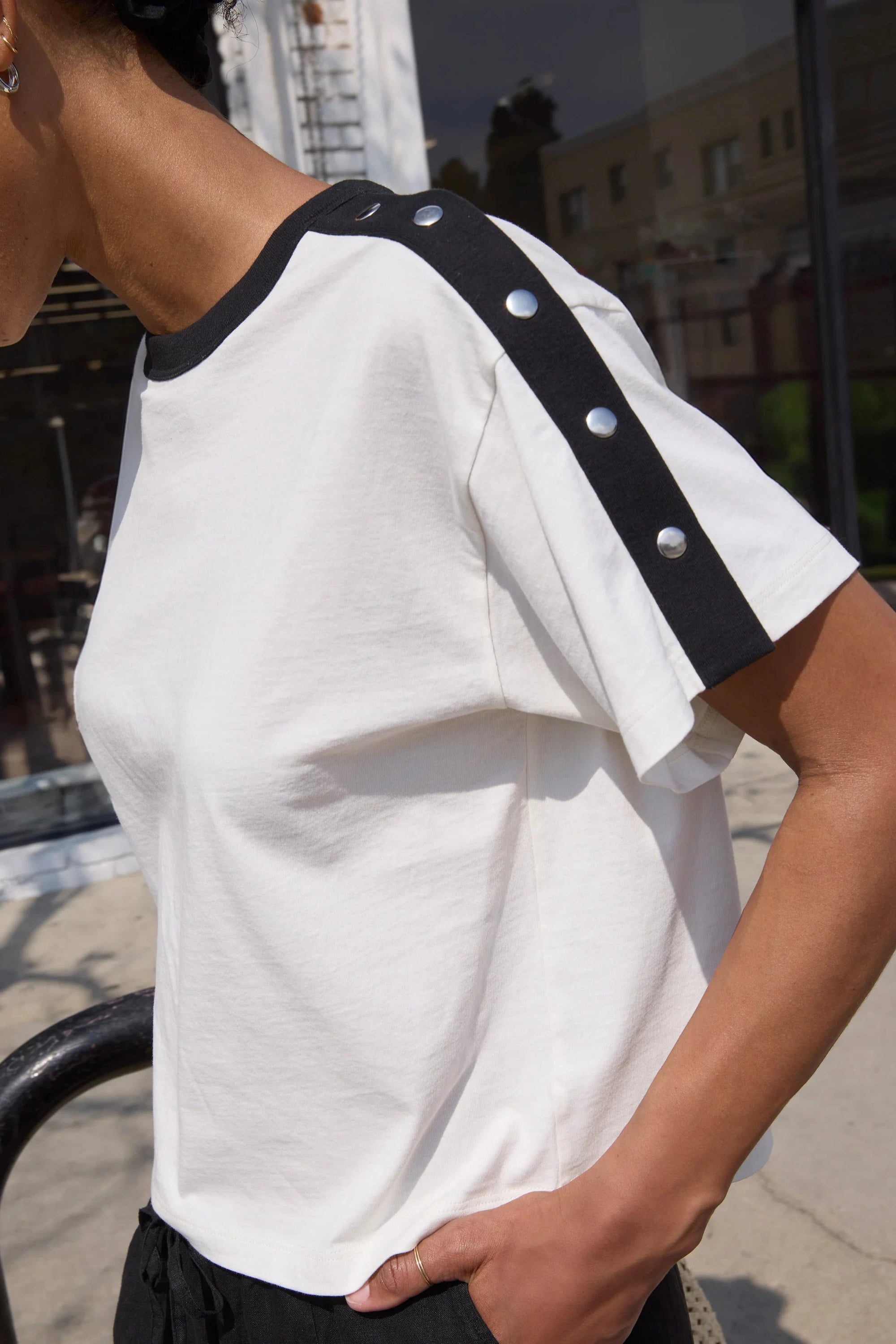 Person wearing a white t-shirt with black trim and buttons outdoors.