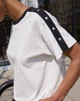 Person wearing a white t-shirt with black trim and buttons outdoors.