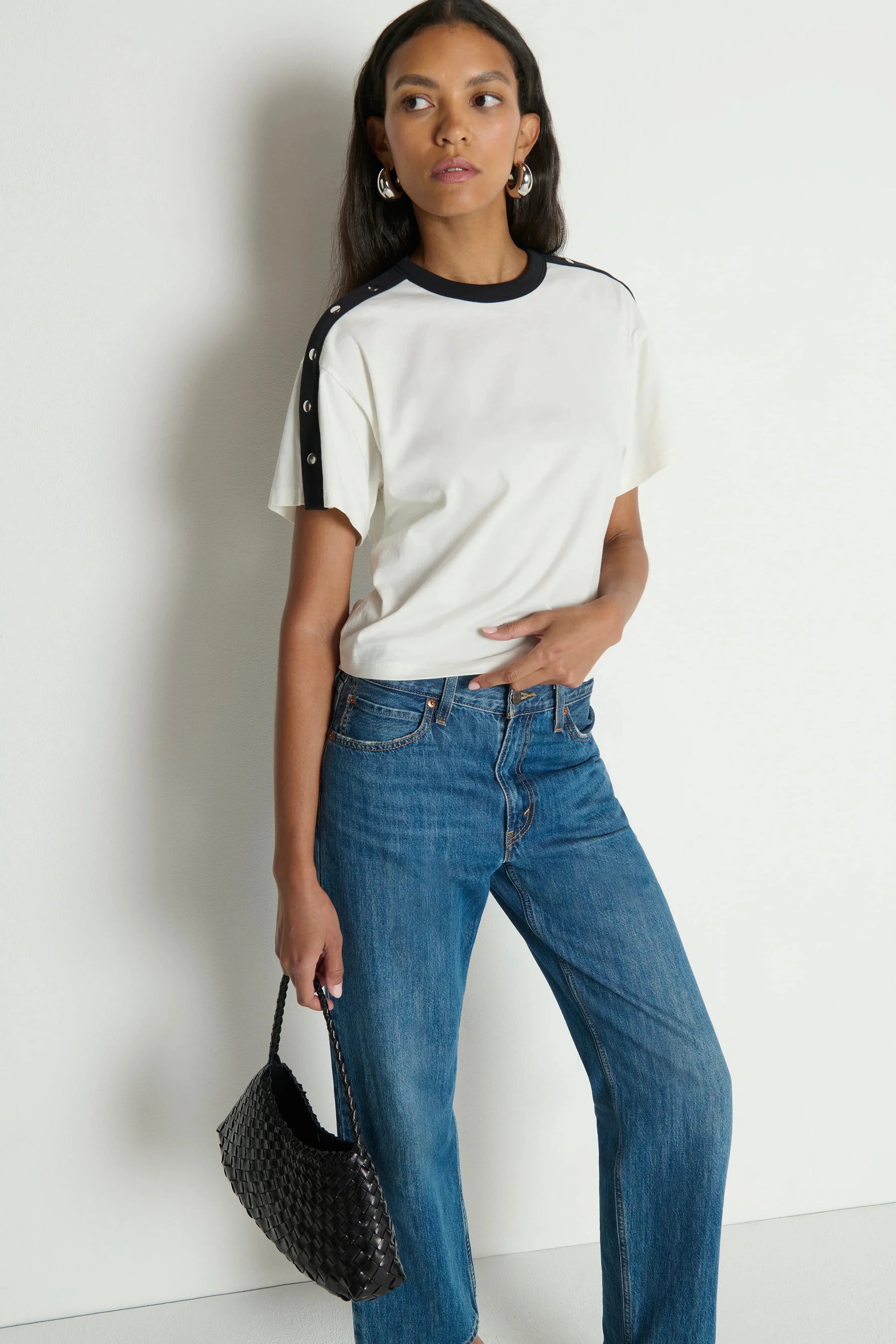 Woman wearing a white t-shirt with black sleeves and blue jeans, holding a black handbag against a plain background.