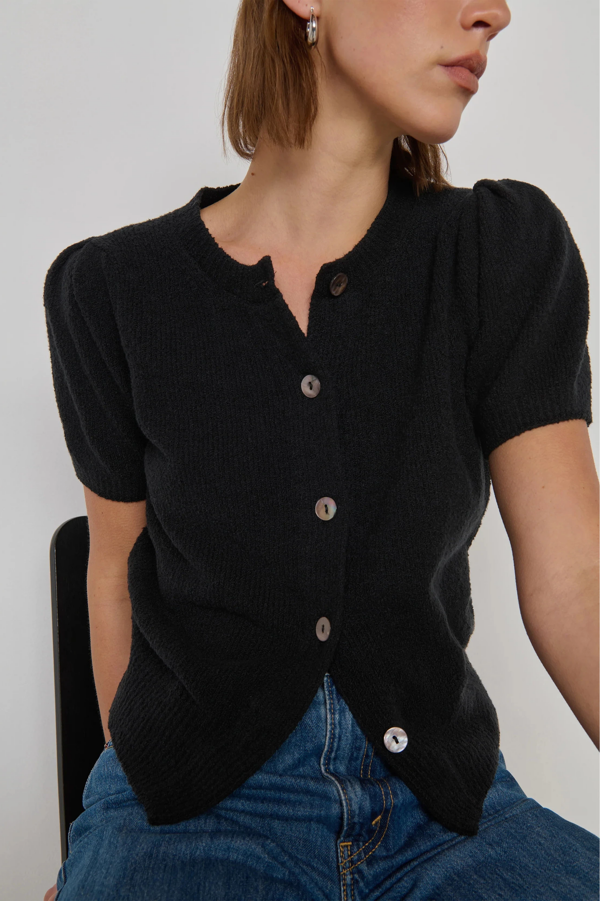 Person wearing a black cardigan with short sleeves and buttons, sitting against a plain background.