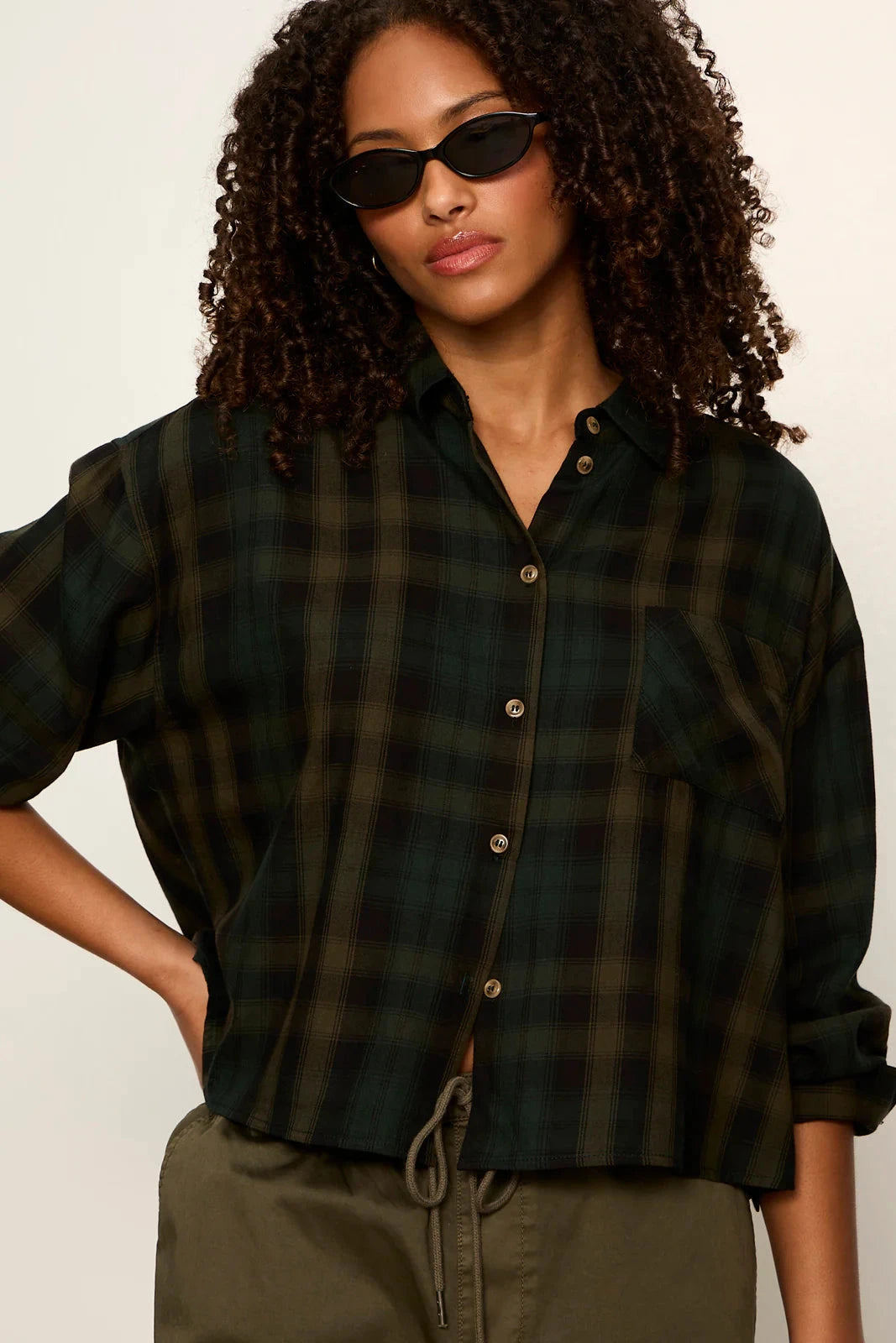 A woman wearing a dark green and black plaid button-up shirt with rolled sleeves, styled with olive green drawstring pants and black sunglasses against a plain off-white background.