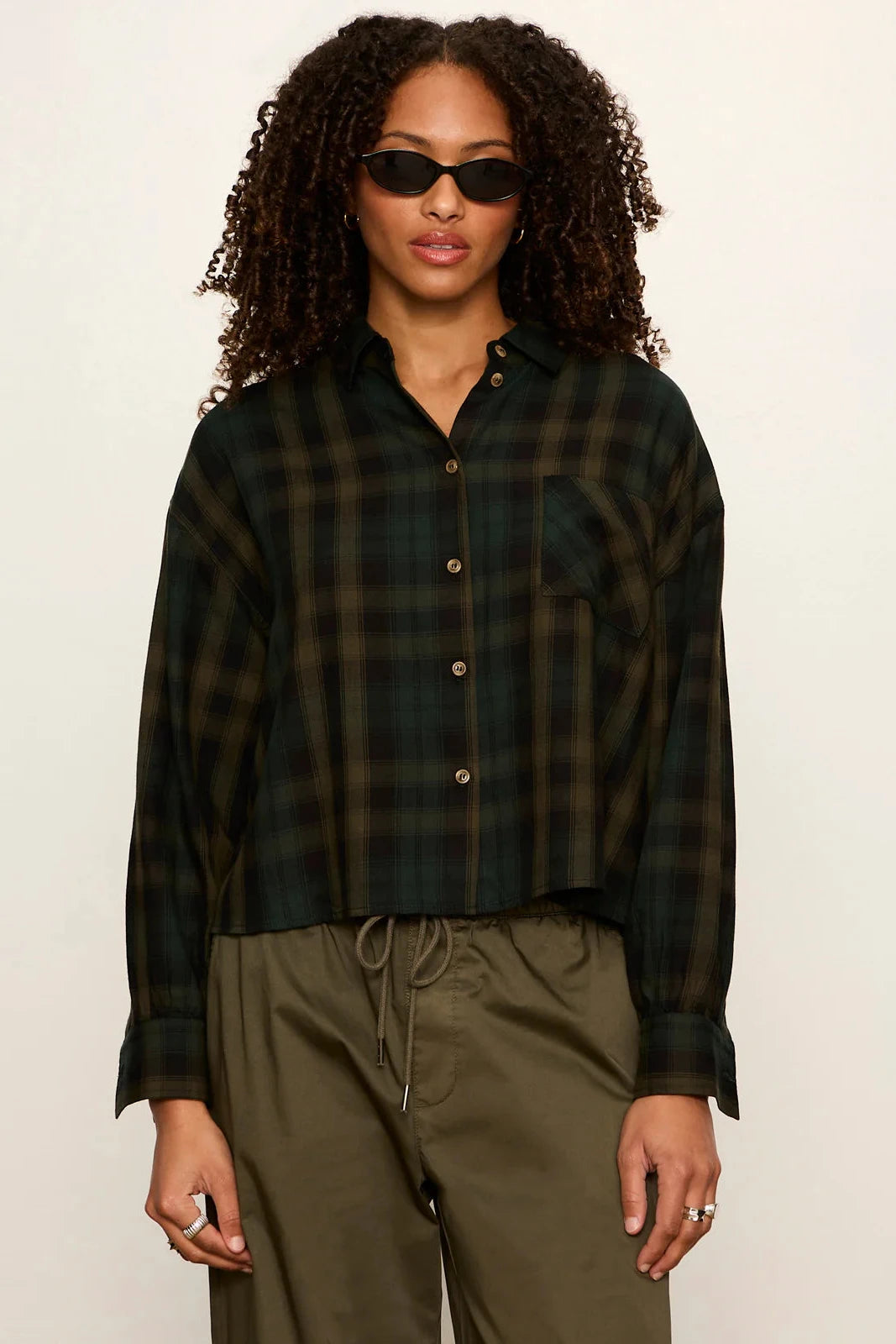 A model wearing a dark green and black plaid button-down shirt with olive green drawstring pants, styled with black sunglasses against a plain off-white background.