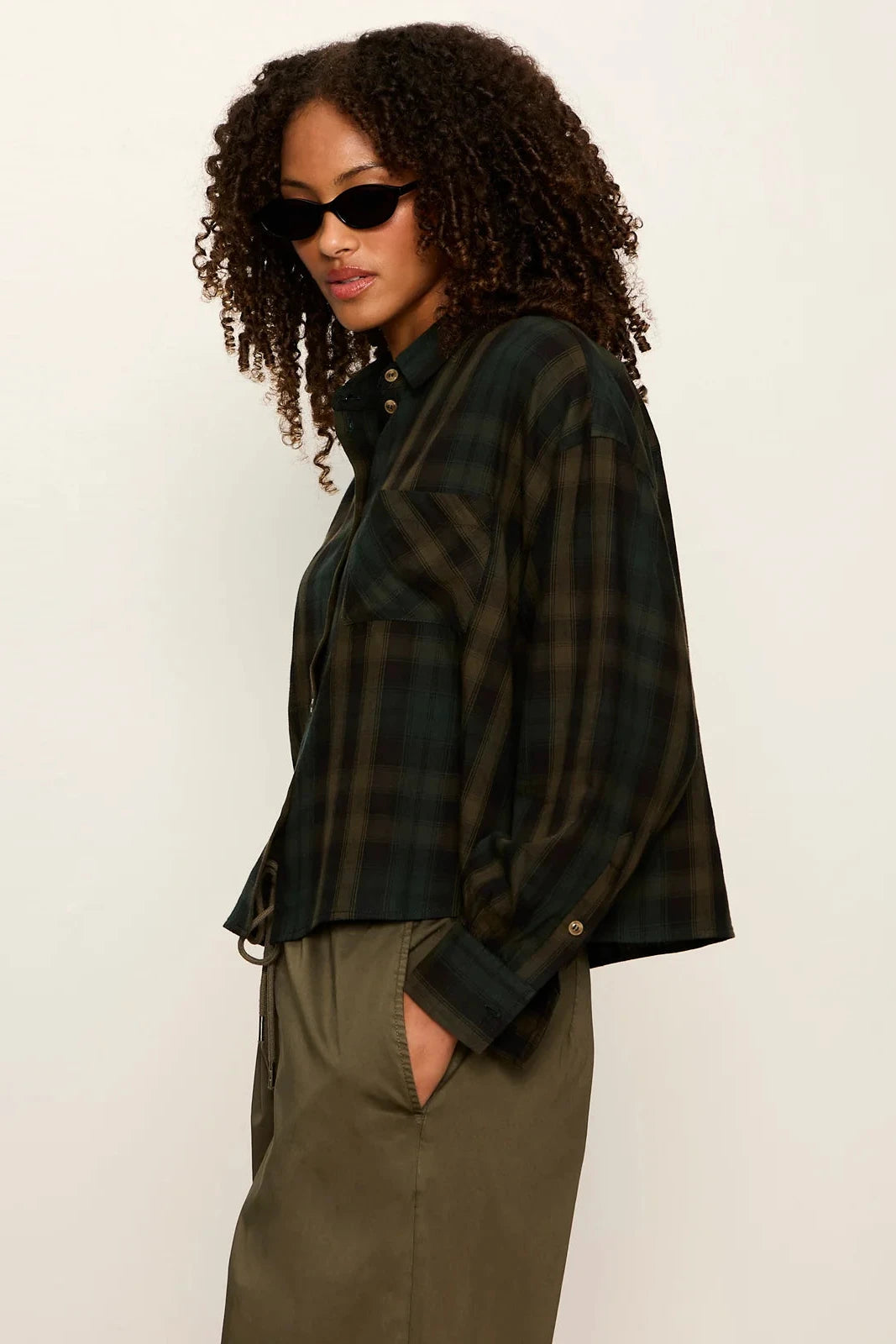 A woman with curly hair wearing a dark green and black plaid button-up shirt and olive green pants, posed against a plain off-white studio background.