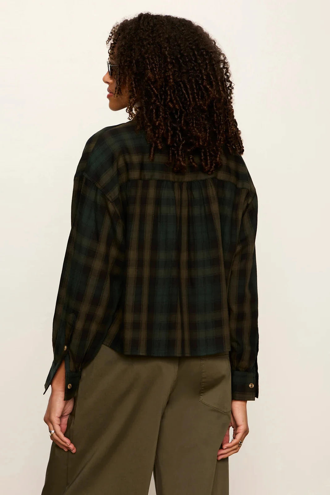 A woman shown from the back wearing a dark green and black plaid flannel shirt and olive green pants, photographed against a plain off-white studio background.