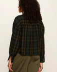 A woman shown from the back wearing a dark green and black plaid flannel shirt and olive green pants, photographed against a plain off-white studio background.