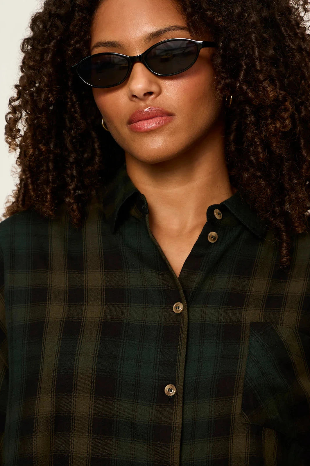 A close-up of a model wearing a dark green and black plaid button-down shirt with brown buttons, styled with black sunglasses.