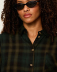 A close-up of a model wearing a dark green and black plaid button-down shirt with brown buttons, styled with black sunglasses.
