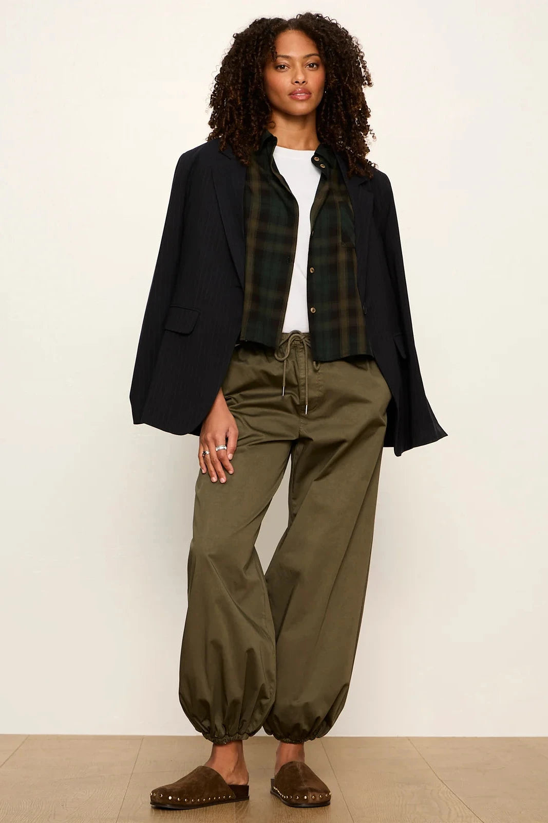 A model stands against a plain off-white studio wall wearing a dark navy blazer, a green and black plaid shirt, a white top, olive green drawstring pants, and brown slip-on shoes.