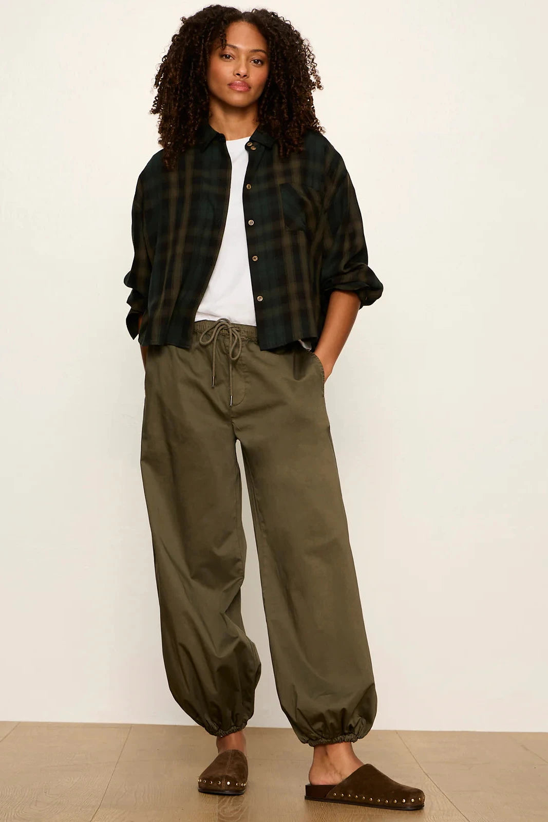 A woman stands against a plain off-white wall wearing a dark green and black plaid button-up shirt over a white t-shirt, olive green baggy drawstring pants, and brown mule sandals.