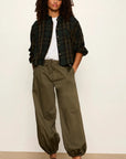 A woman stands against a plain off-white wall wearing a dark green and black plaid button-up shirt over a white t-shirt, olive green baggy drawstring pants, and brown mule sandals.
