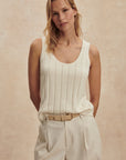 Woman wearing a white sleeveless top and beige pants against a beige background