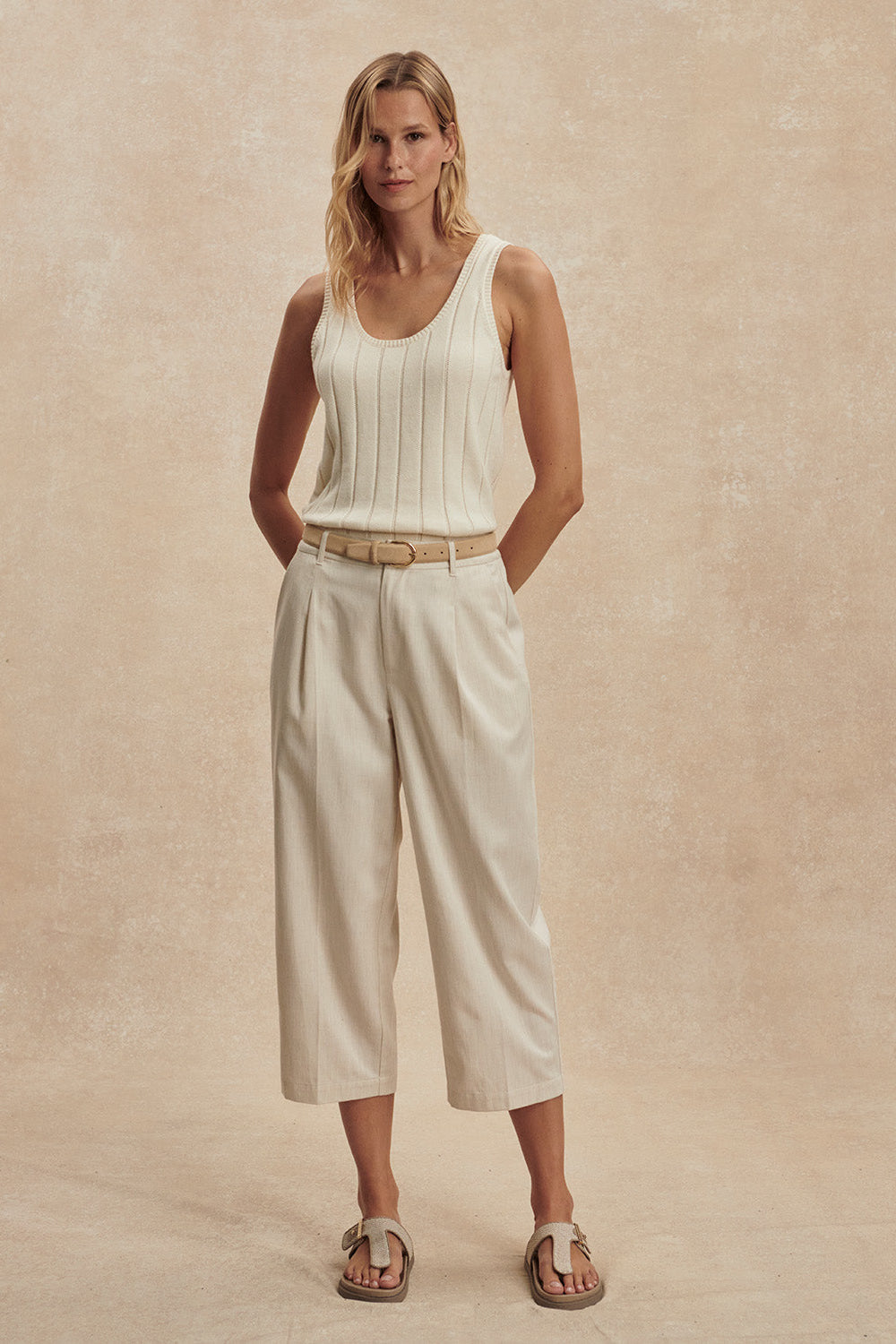 Woman wearing a white sleeveless top and beige pants against a beige background