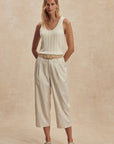 Woman wearing a white sleeveless top and beige pants against a beige background