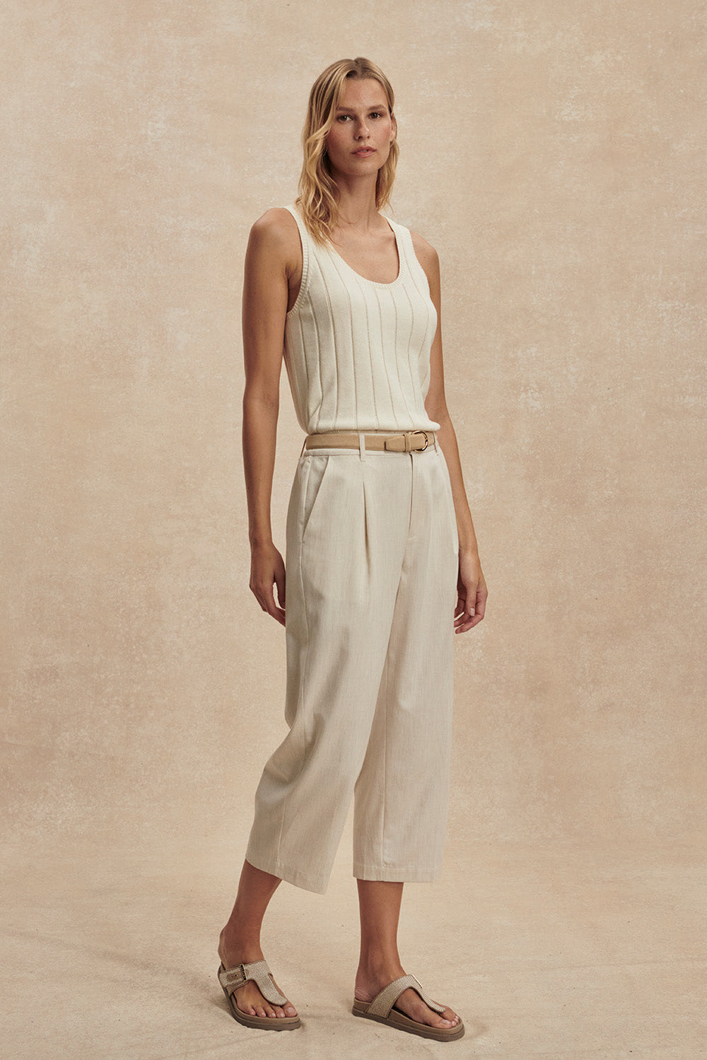 Woman wearing a white sleeveless top and beige pants against a beige background