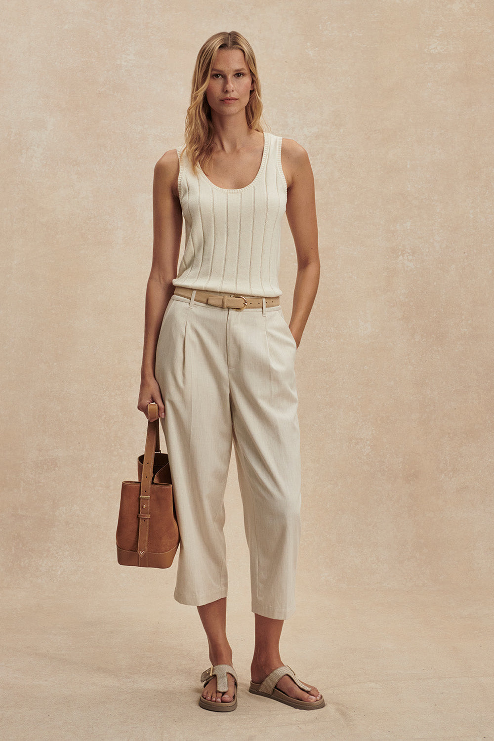 Woman wearing a white sleeveless top and beige pants, holding a brown leather bag on a beige background