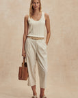 Woman wearing a white sleeveless top and beige pants, holding a brown leather bag on a beige background