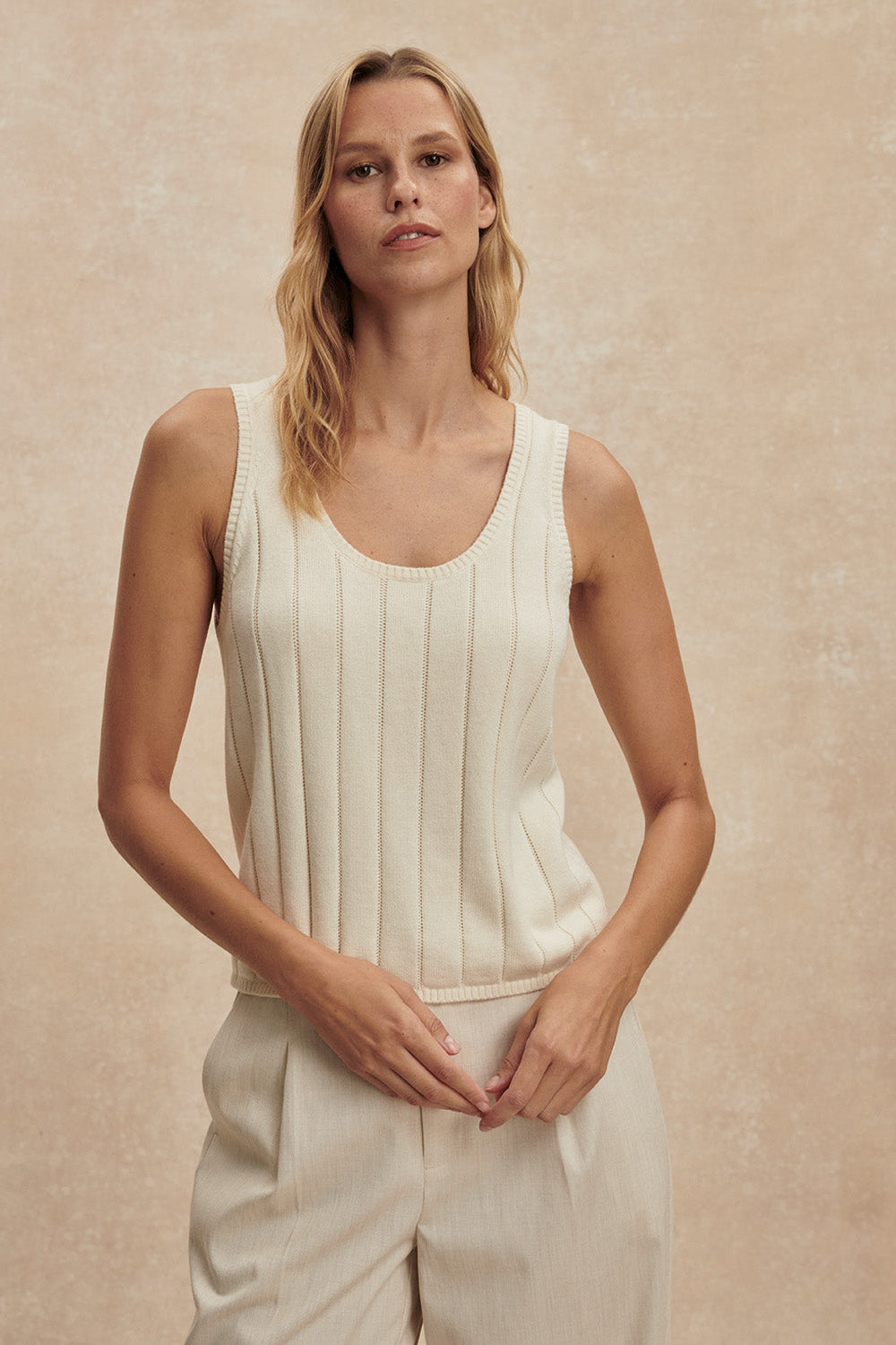 Woman wearing a white sleeveless top and pants against a beige background