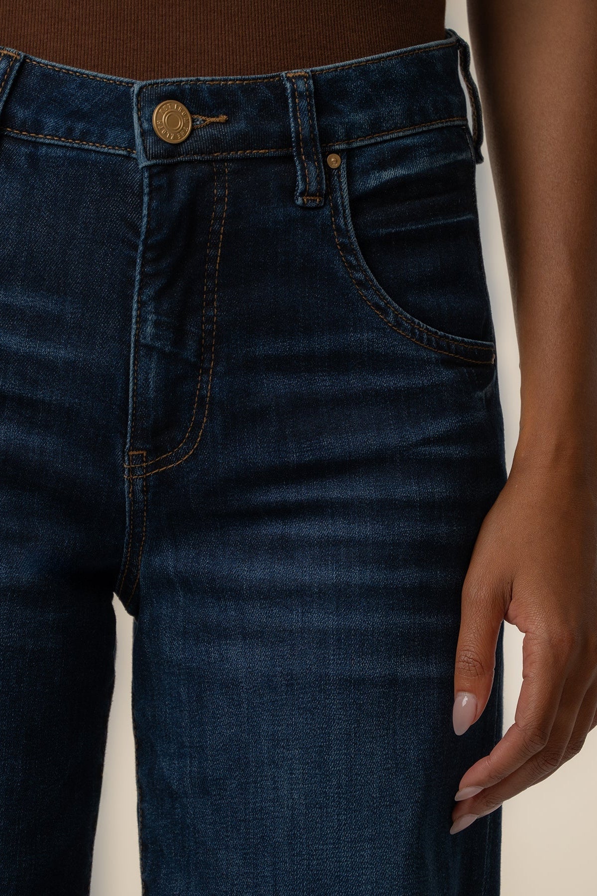 Close-up of a model wearing dark blue denim jeans with a brown top, showing the waistband, button, pocket, and hand.