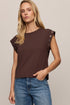 A woman wearing a dark brown short-sleeve top with decorative eyelet details on the shoulders and blue jeans, photographed against a plain beige studio background.