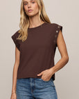 A woman wearing a dark brown short-sleeve top with decorative eyelet details on the shoulders and blue jeans, photographed against a plain beige studio background.