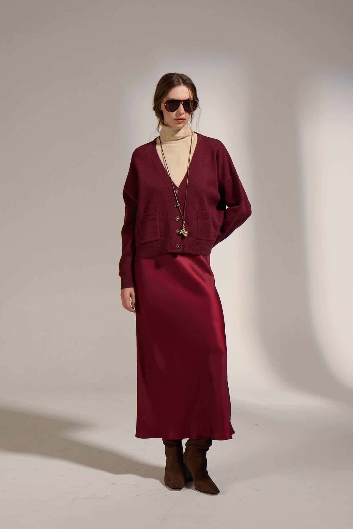 A model stands against a warm beige studio backdrop wearing a matching burgundy cardigan and midi skirt set, a cream turtleneck, layered gold necklaces, sunglasses, and brown suede boots.