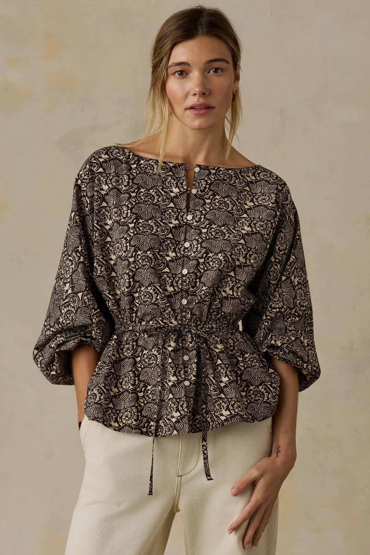 A woman wearing a dark brown and beige snakeskin print peplum blouse with three-quarter sleeves and cream pants, standing against a textured beige wall.