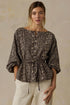 A woman wearing a dark brown and beige snakeskin print peplum blouse with three-quarter sleeves and cream pants, standing against a textured beige wall.