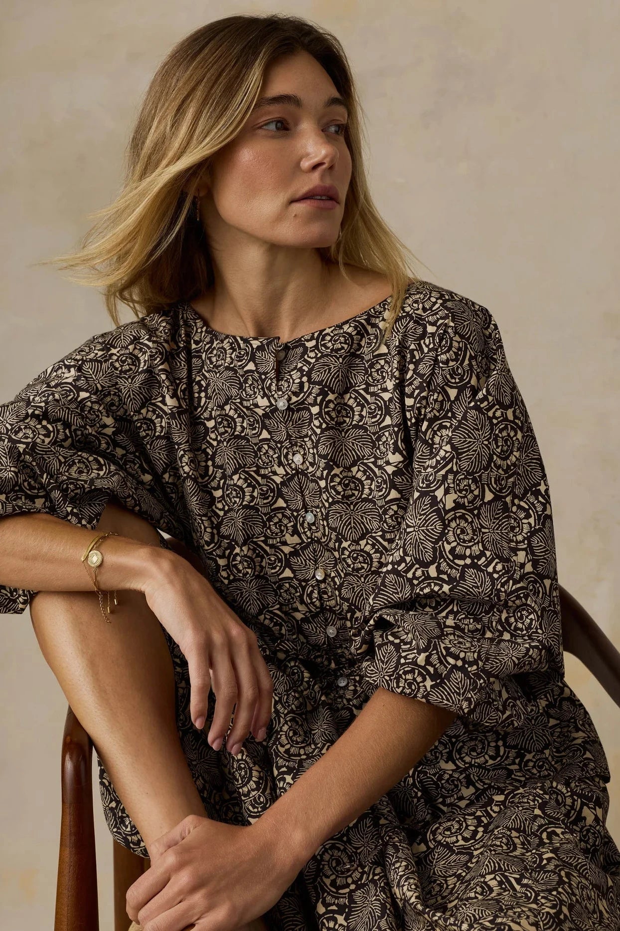 Woman wearing a patterned dress sitting on a chair against a beige background