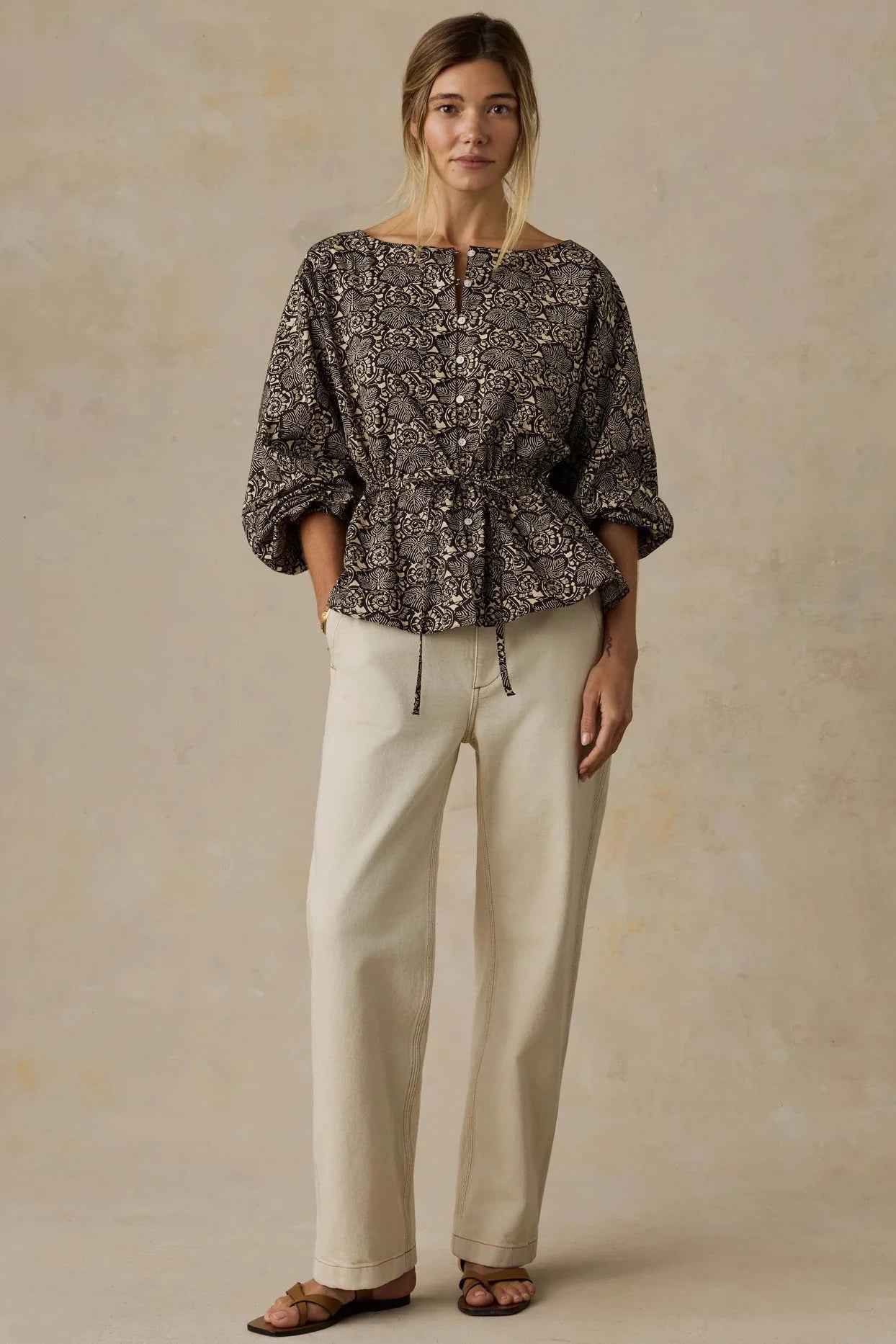Woman wearing a patterned top and beige pants on a beige background