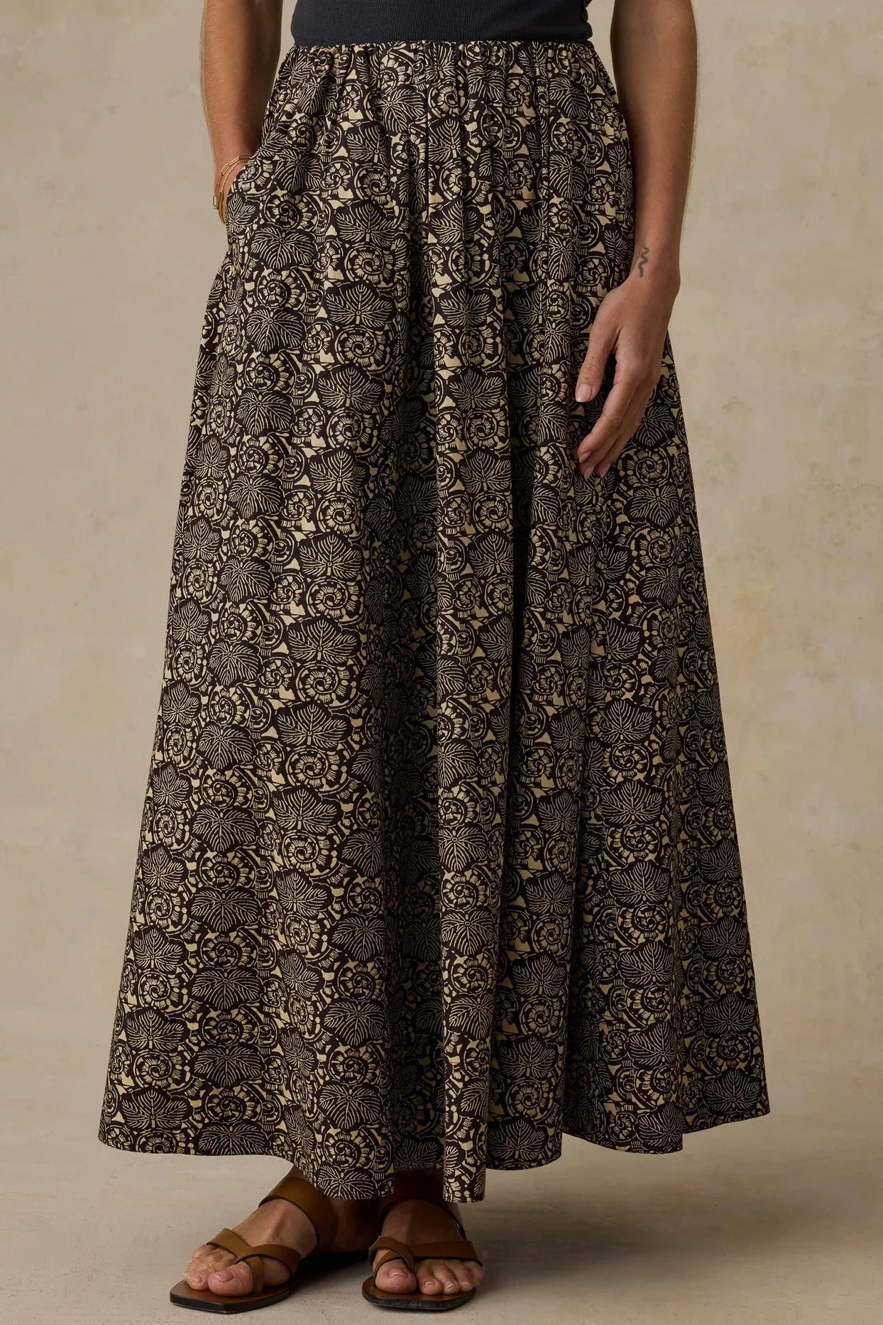 A high-waisted, full-length maxi skirt with an intricate dark brown and beige floral jacquard pattern, shown on a model wearing brown leather sandals against a textured beige studio backdrop.
