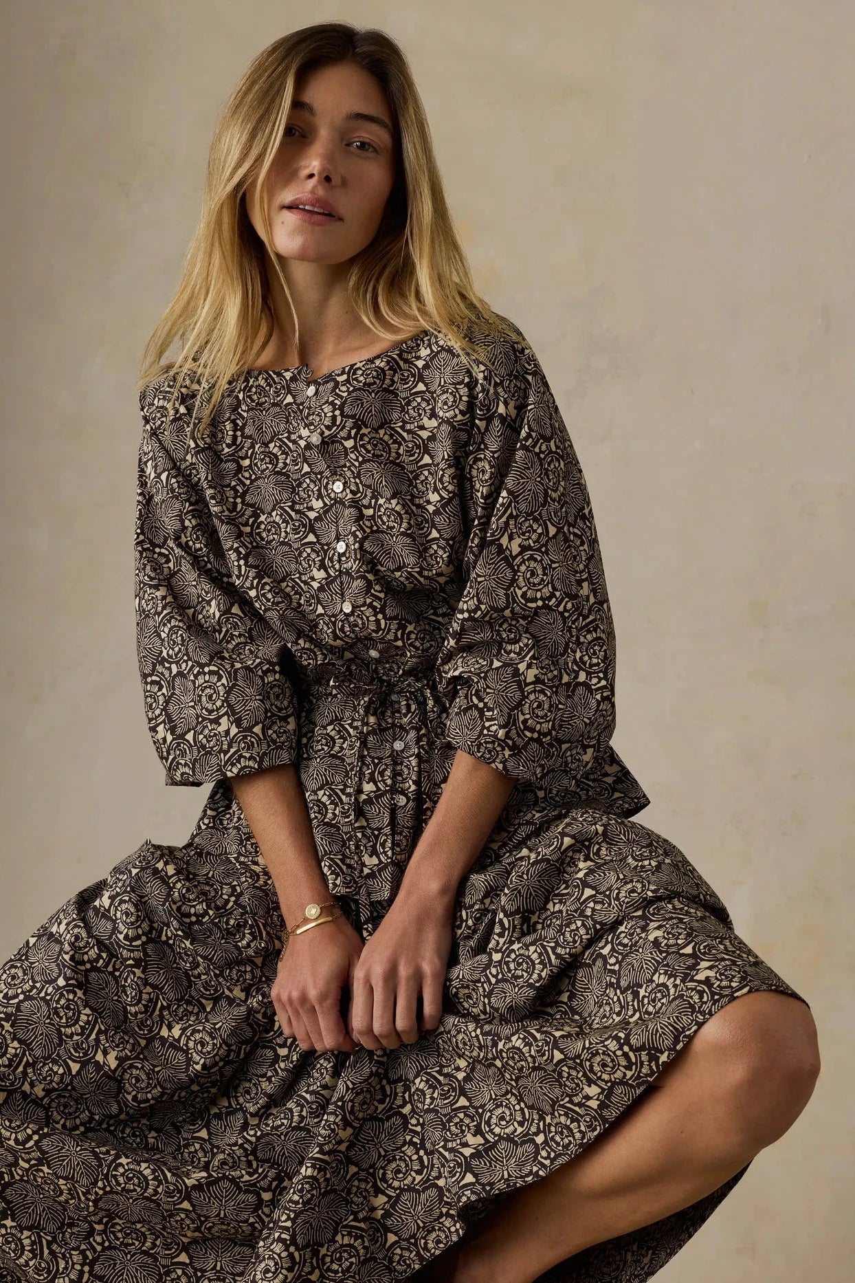 A woman wearing a long-sleeved midi dress with a dark brown and beige floral print, sitting against a neutral textured beige background.