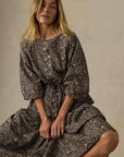 A woman wearing a long-sleeved midi dress with a dark brown and beige floral print, sitting against a neutral textured beige background.