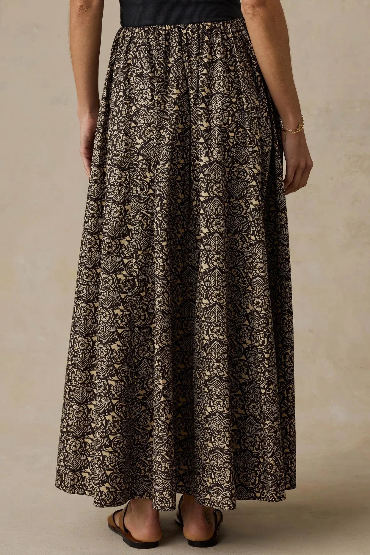 A woman wearing a long, full-length skirt with an intricate black, brown, and beige floral or lace-like pattern, paired with a black top and flat sandals, shown from the back against a neutral beige background.