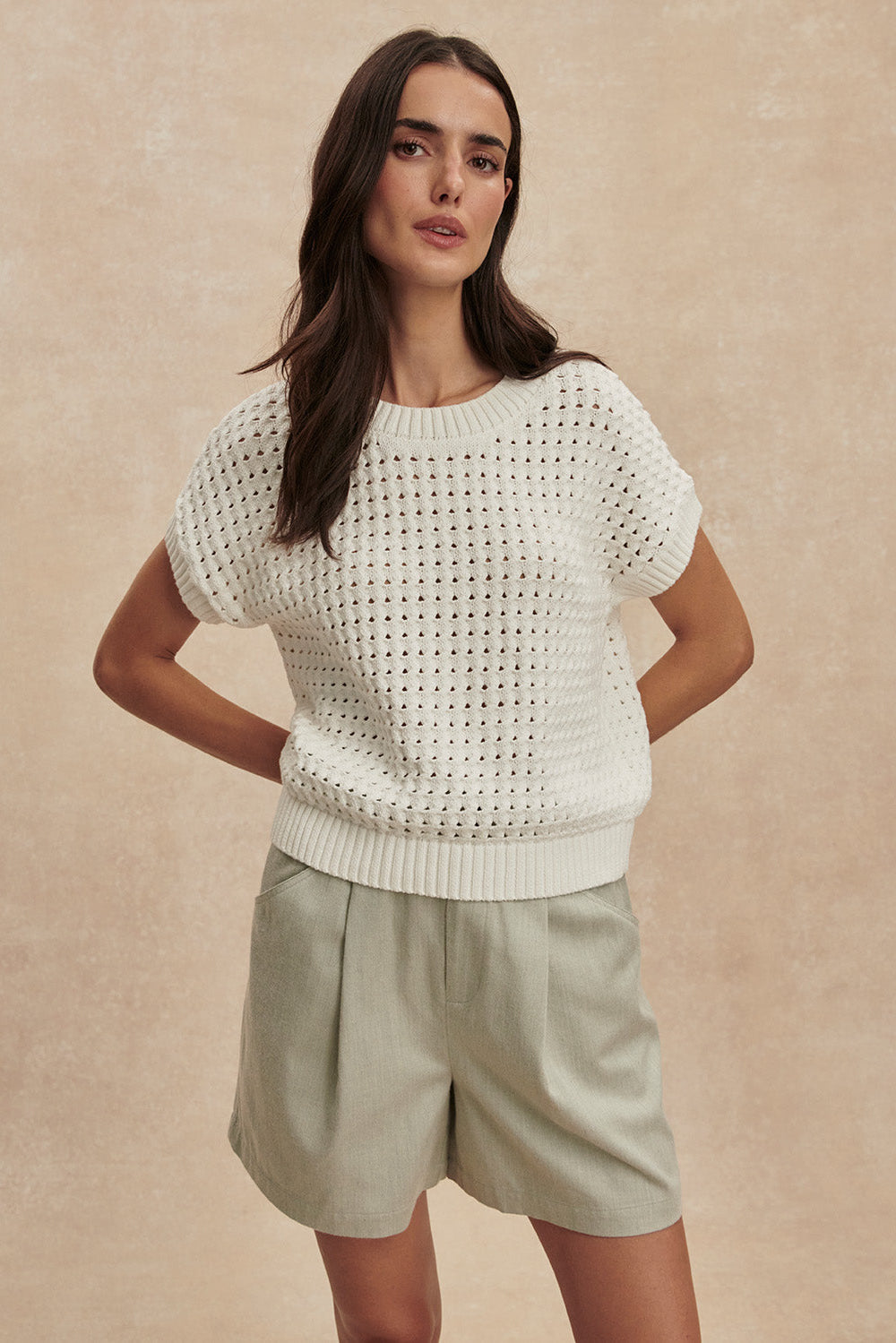 Woman wearing a white knitted sweater and light green shorts against a beige background