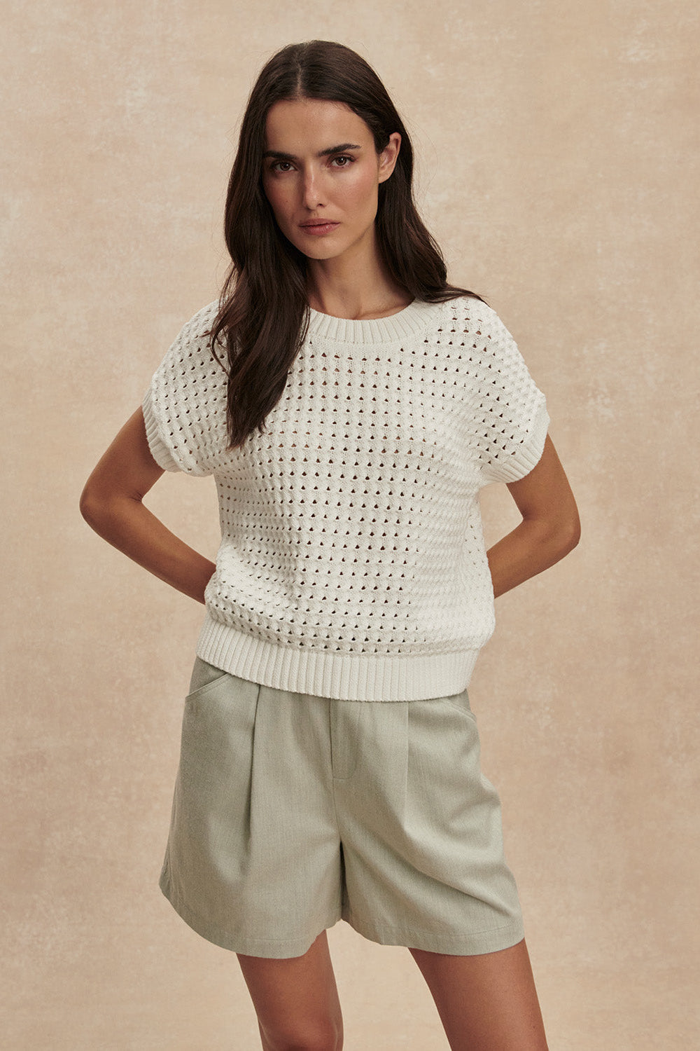 Woman wearing a white knitted top and beige shorts against a beige background