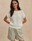 Woman wearing a white knitted top and beige shorts against a beige background