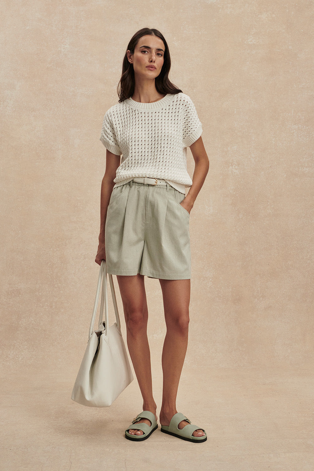 Woman wearing a white textured top and light green shorts, holding a beige bag on a beige background
