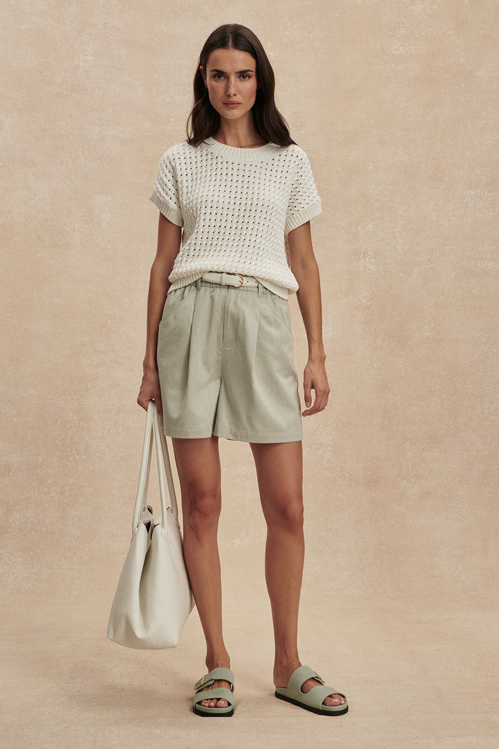 Woman wearing a white textured top and light green shorts, holding a matching tote bag on a beige background.