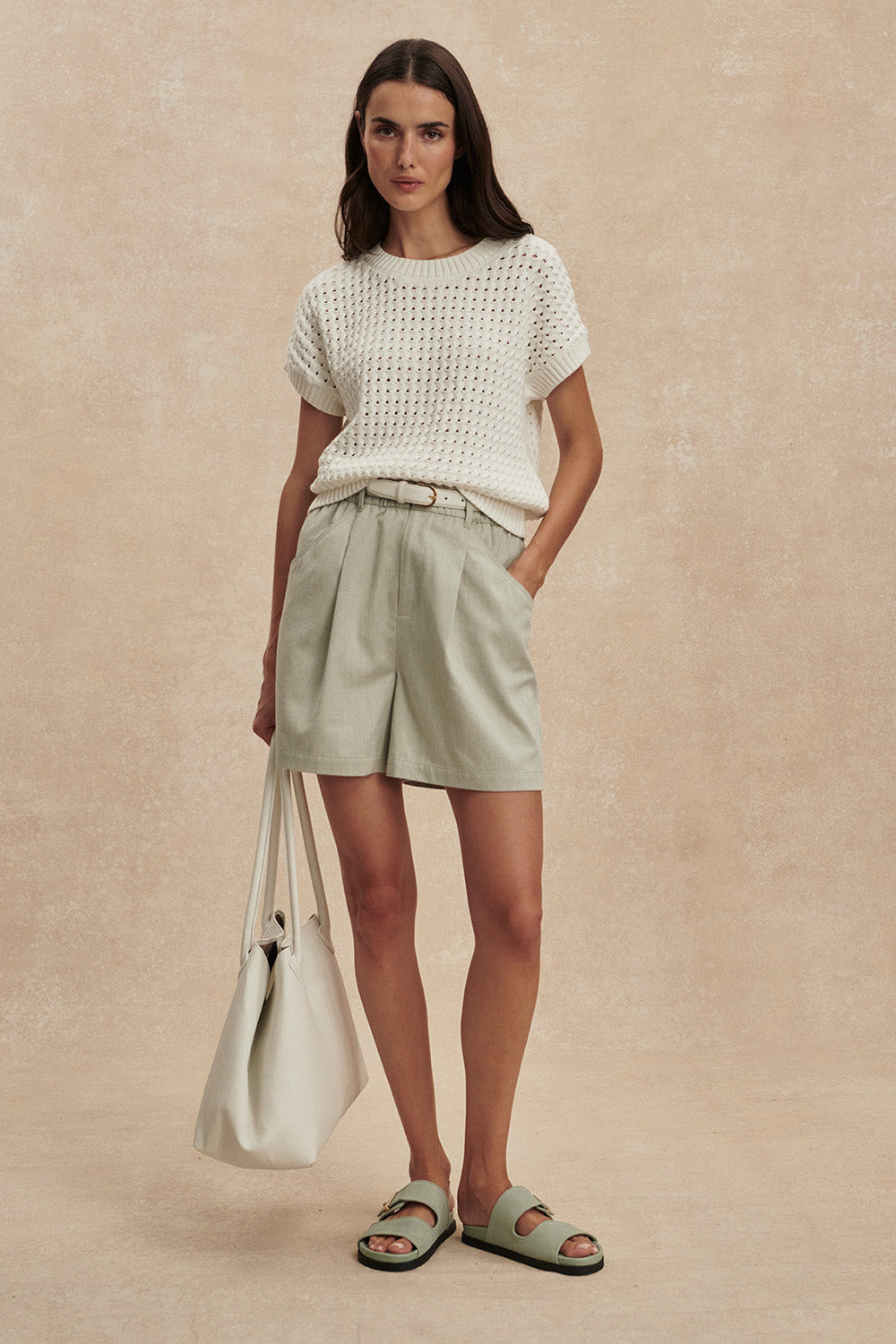 Woman wearing a white textured top and light green shorts, standing against a beige background.