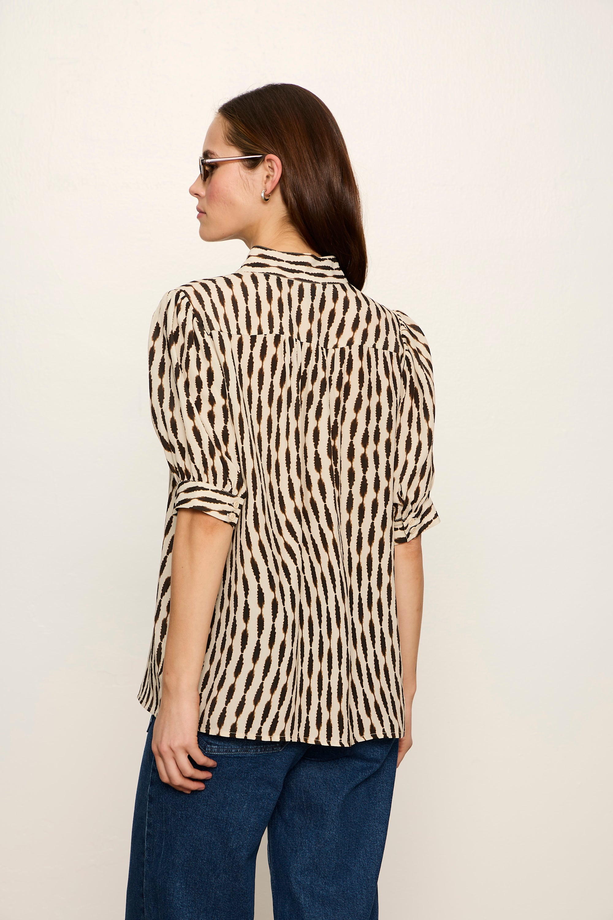 Woman wearing a patterned blouse and blue pants against a plain background
