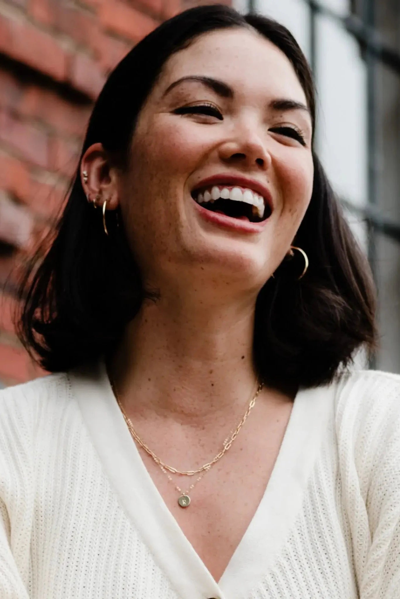 Woman laughing outdoors with a brick wall and window in the background