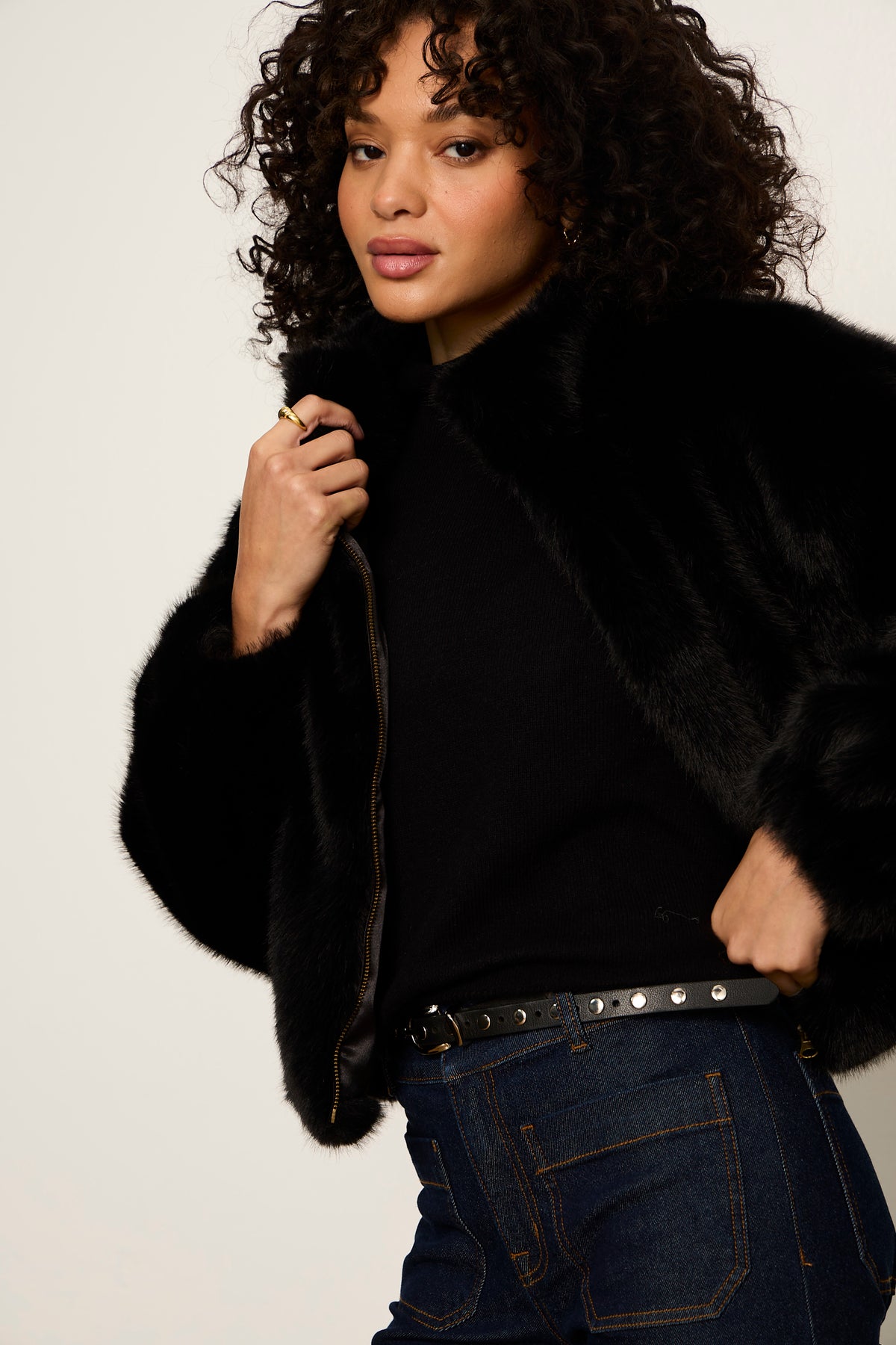 Woman wearing a black fur coat and dark jeans against a plain background