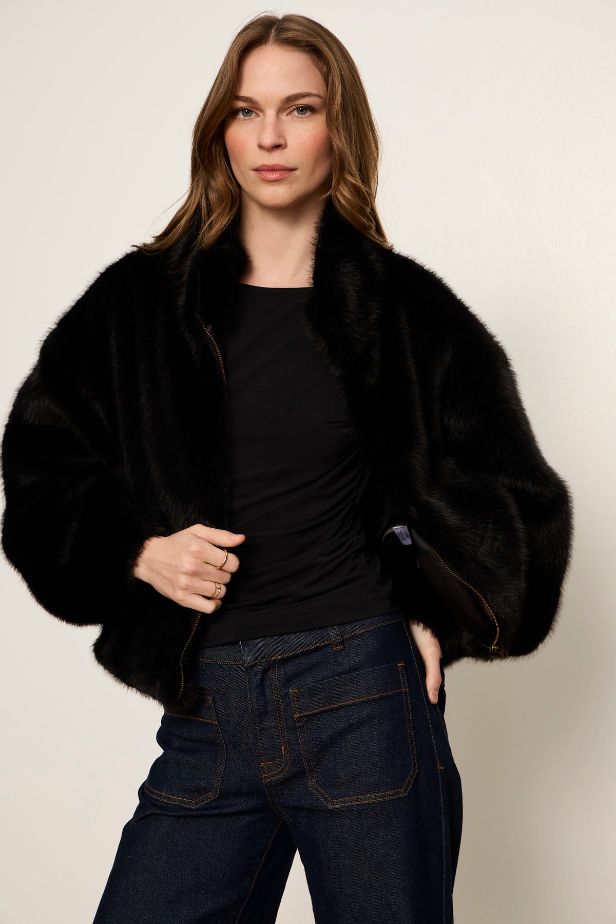 Woman wearing a black fur coat over a black top and blue jeans on a plain background