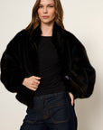 Woman wearing a black fur coat over a black top and blue jeans on a plain background