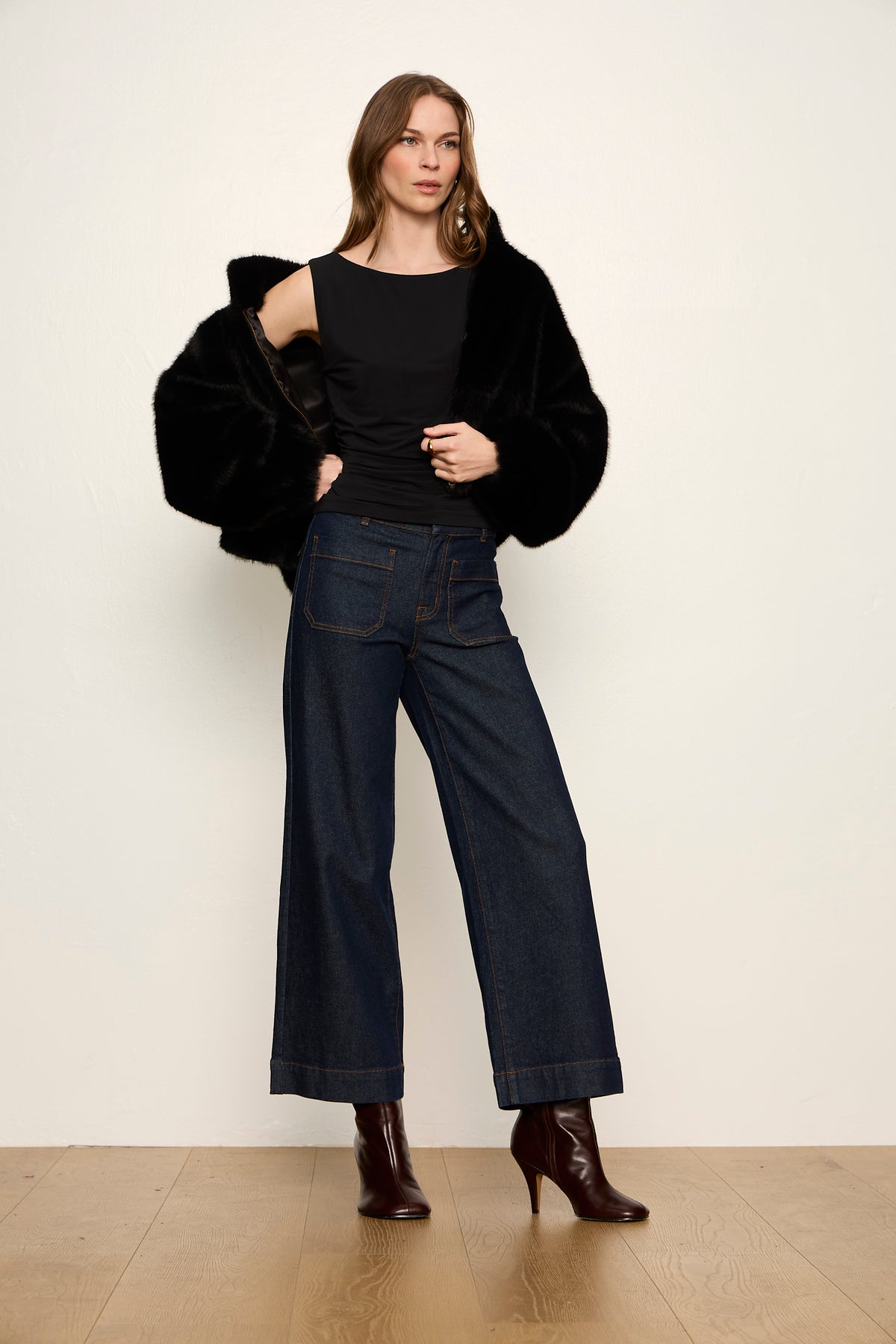 Woman wearing a black fur coat, black top, and dark jeans on a white background