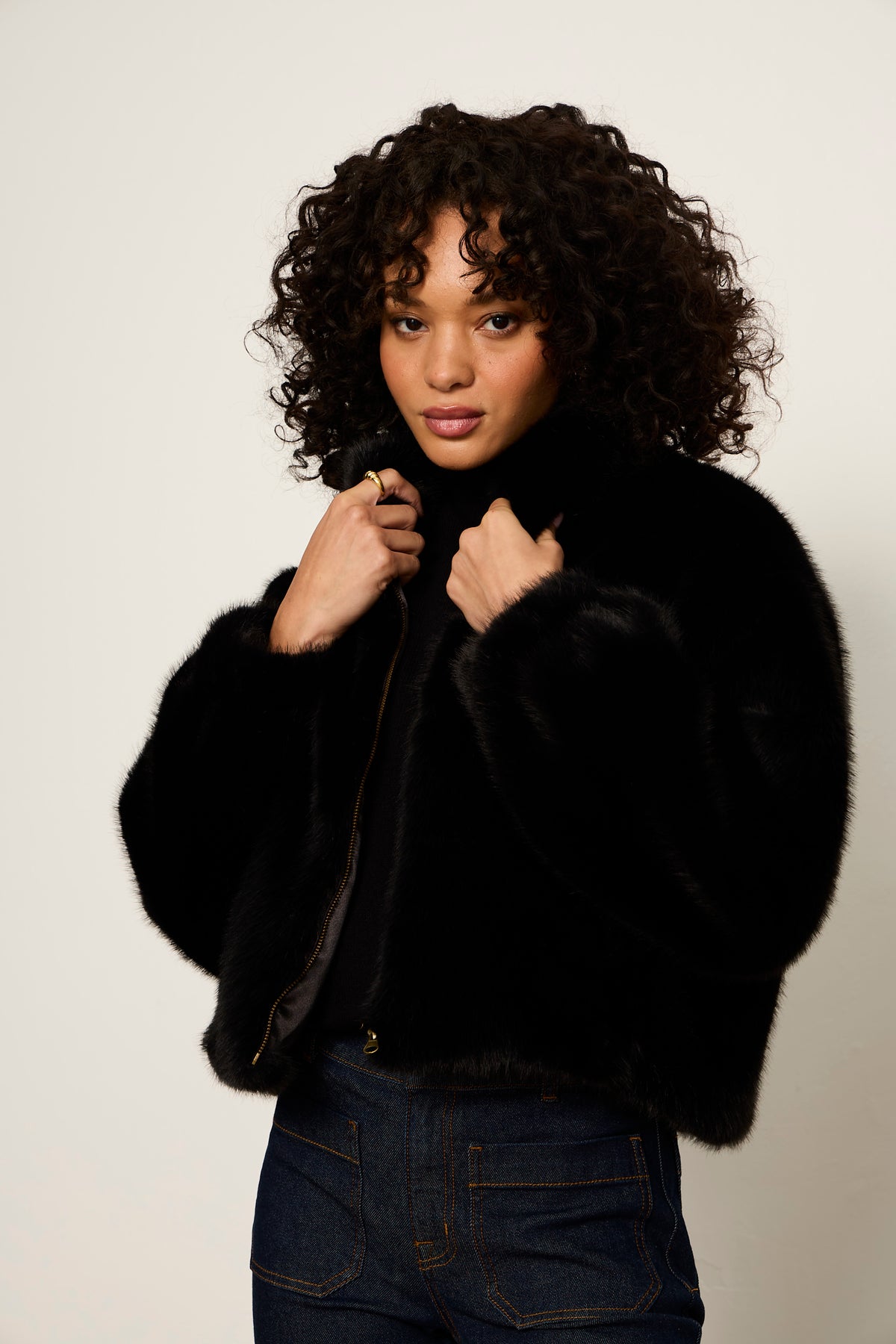A model with curly dark hair wearing a black cropped faux fur jacket over a black top and dark blue jeans, posing against a light gray studio background.