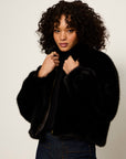 A model with curly dark hair wearing a black cropped faux fur jacket over a black top and dark blue jeans, posing against a light gray studio background.