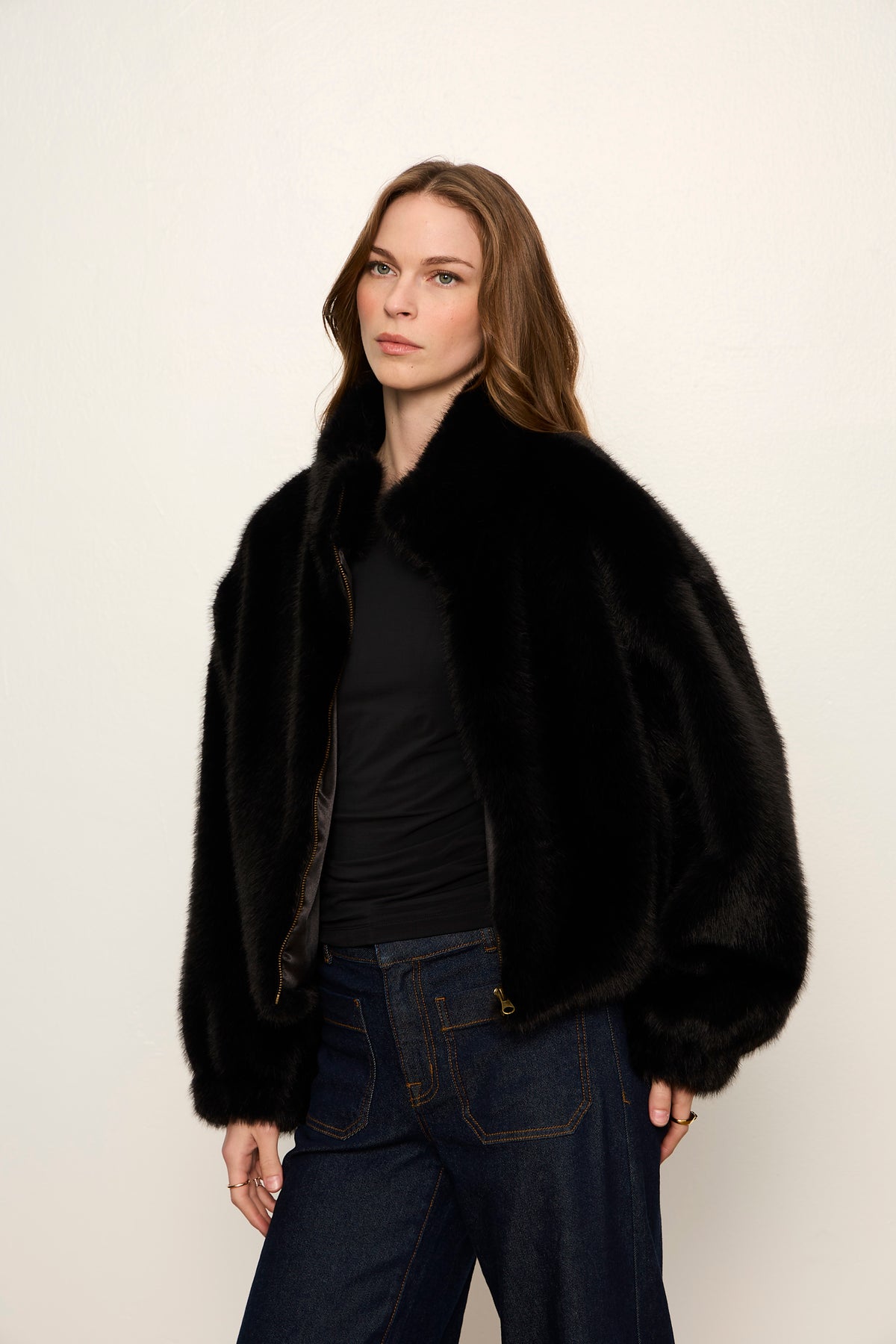 Woman wearing a black fur coat and dark jeans against a plain background