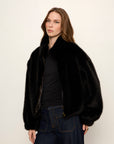 Woman wearing a black fur coat and dark jeans against a plain background